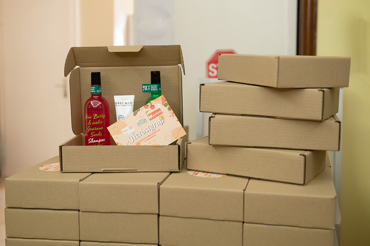 Geöffnete Spendenbox der Tafel Hessen mit Pflegeprodukten und Herbstgrußkarte, umgeben von gestapelten Versandkartons im Rahmen der Deutschen Postcode Lotterie Herbstaktion.