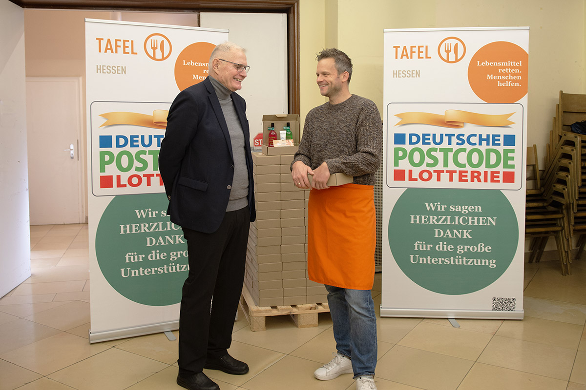 Zwei Personen im Gespräch vor Bannern der Tafel Hessen und Deutschen Postcode Lotterie, daneben gestapelte Spendenboxen als Teil der Herbstaktion.